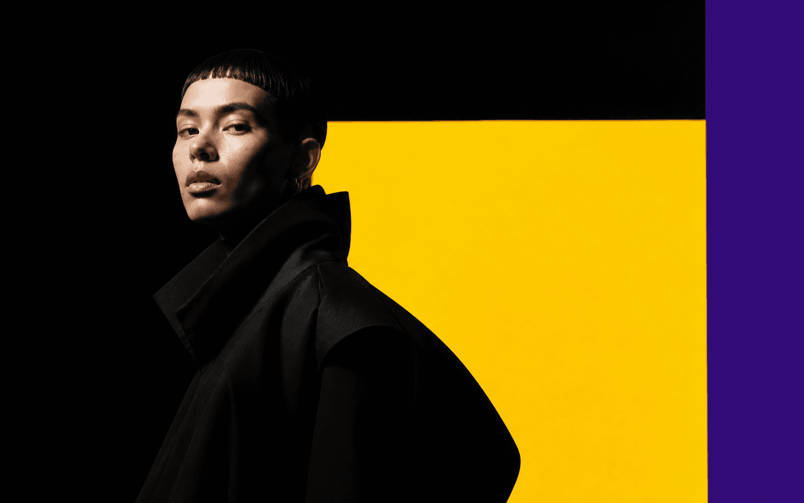 Fictional synthetic editorial creator in architectural black clothing against yellow and purple studio panels.
